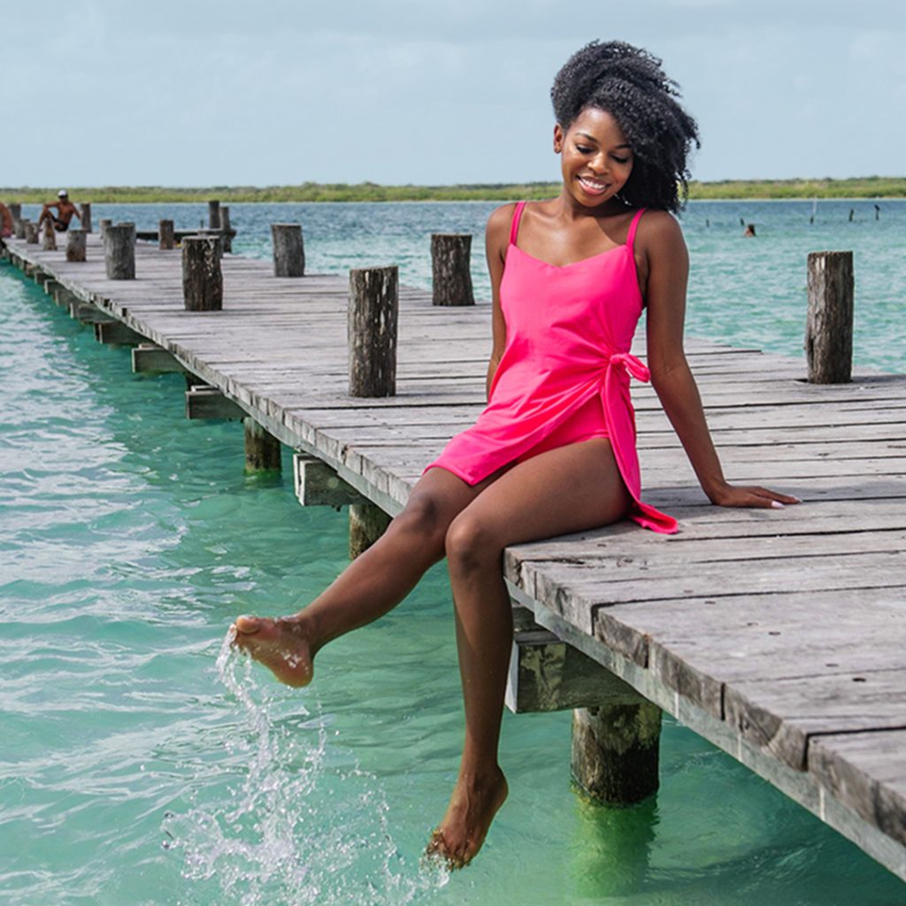 Young African American model sits on the edge of a dock wearing a hot pink swim dress. The slit shows the underlayer of the suit. 