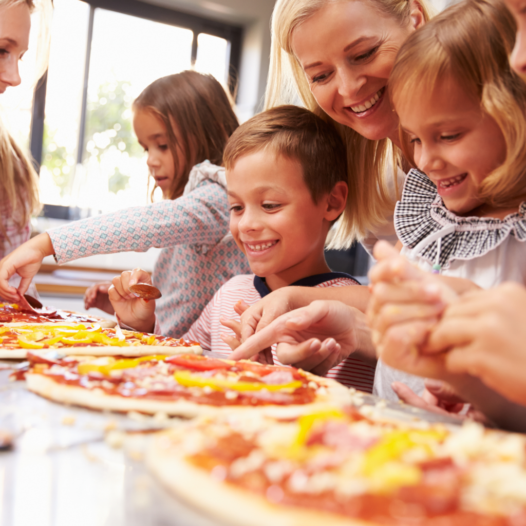 A family of smiling children and mother build pizzas. 